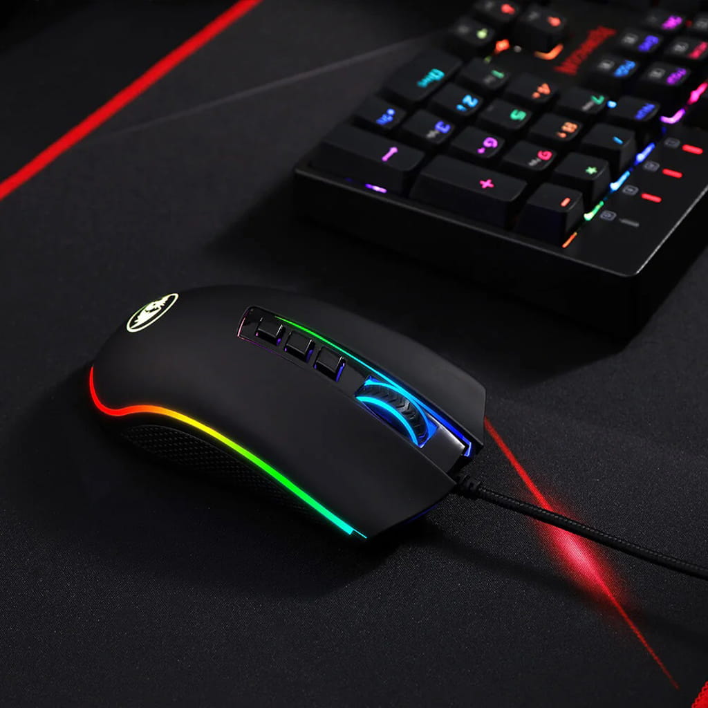 Left side view of the black Redragon M711 FPS gaming mouse with illuminated RGB lighting, resting on a mousepad next to a keyboard